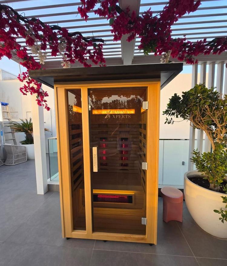 Outdoor infrared sauna installed under a pergola with SaunaXperts logo etched on the glass door